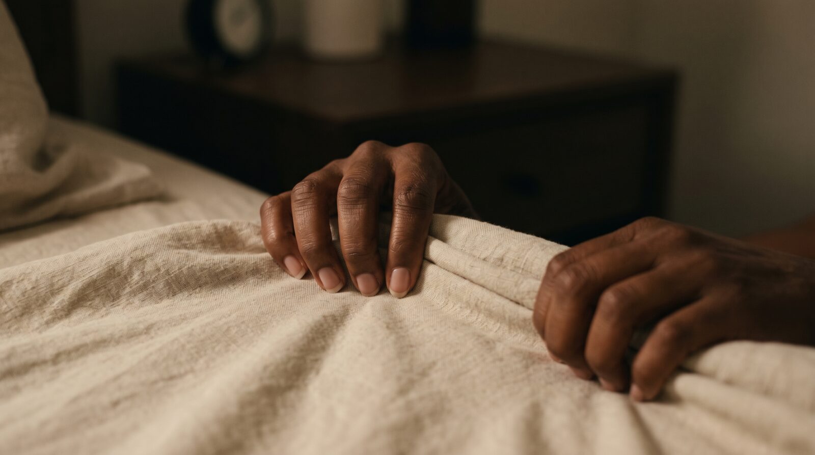 A close up view of a Black woman's hands gripping linen bed sheets in dim early morning light.